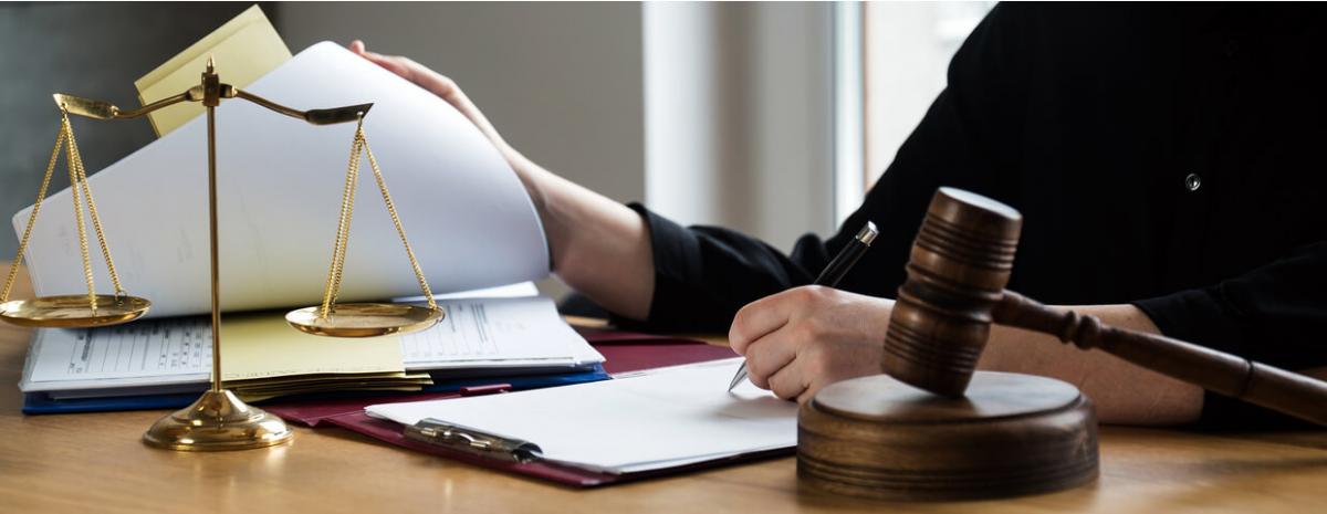 Lawyer working at desk flipping through and signing paperwork with judge gavel and scales of justice on desk.