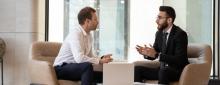 two male business partners sitting in chairs, talking about business or services with a laptop on a table