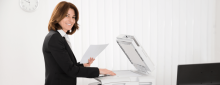 Business woman smiling while holding stack of papers in front of printer machine.